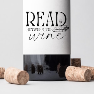 May include: A dark wine bottle with a white label featuring the text "READ BETWEEN THE wine". The label also includes a small illustration of a wine glass and books. Several corks are scattered around the bottle.