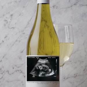 May include: A bottle of white wine with a custom label featuring an ultrasound image. The label reads "Pour some bubbly, there's gonna be a baby!" and includes the baby's name and due date. A wine glass filled with wine is next to the bottle.