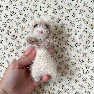 May include: A handmade crochet doll with a white body and a brown and white head. The doll has black eyes and a pink mouth. The doll is held against a floral patterned background.