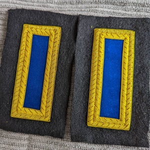 May include: Two blue fabric patches with gold trim, sewn onto a black fabric background. The patches are rectangular and have a gold border with a braided design.