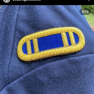 May include: A gold-colored, embroidered insignia with a blue center, likely a military rank or emblem. The insignia is oval-shaped with a detailed, textured border and two vertical gold stripes. It is set against a dark blue fabric background.