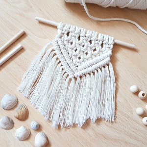 May include: A white macrame wall hanging with a triangular pattern and fringe. The hanging is made with natural cotton cord and is attached to a wooden dowel.