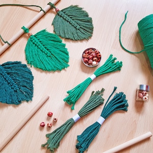 Könnte beinhalten: Grüne Makramee-Feder-Wandbehänge mit Holzstab, grünem Garn und braunen Holzperlen.