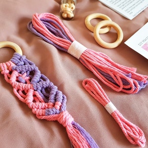 Op de afbeelding: Een roze en paarse macramé wandhanger met een houten ring en een kwast. De macramé is gemaakt met twee strengen garen, een roze en een paarse. De wandhanger is een doe-het-zelfproject.