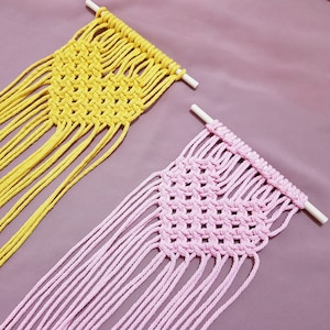 Op de afbeelding: Twee macramé wandhangers, een gele en een roze, met franjes aan de onderkant. De hangers zijn gemaakt met een eenvoudig knoopmotief en zijn bevestigd aan houten latten.