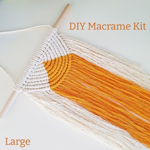 Op de afbeelding: DIY Macramee-set - Groot. Een witte en gele macramee wandhanger met een houten stok aan de bovenkant. De macramee heeft de vorm van een halve cirkel met een franje aan de onderkant.