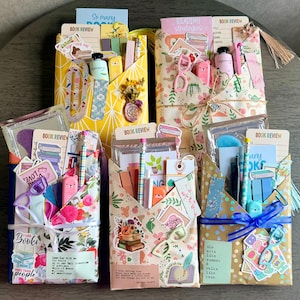May include: Five rectangular gift envelopes wrapped in patterned paper, each containing stationery items like book review cards, pens, stickers, and paper clips. The envelopes feature floral, geometric, and polka dot designs with ribbons.