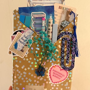 May include: A gold-toned envelope with holographic dots containing stationery items including a book review card, a blue tassel bookmark, a pencil, a pen, stickers, a paperclip, and a sprig of dried teal baby's breath flowers attached to the front.