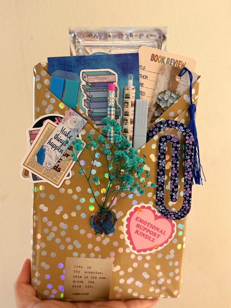 May include: A gold-toned envelope with holographic dots containing stationery items including a book review card, a blue tassel bookmark, a pencil, a pen, stickers, a paperclip, and a sprig of dried teal baby's breath flowers attached to the front.