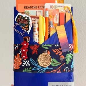 May include: A blue paper-wrapped package containing a reading list, book review card, yellow highlighter, orange pencil, drink mix packet, and a sunset-themed bookmark. It features a floral ribbon, a wax seal, and a label with the text be easy to love.