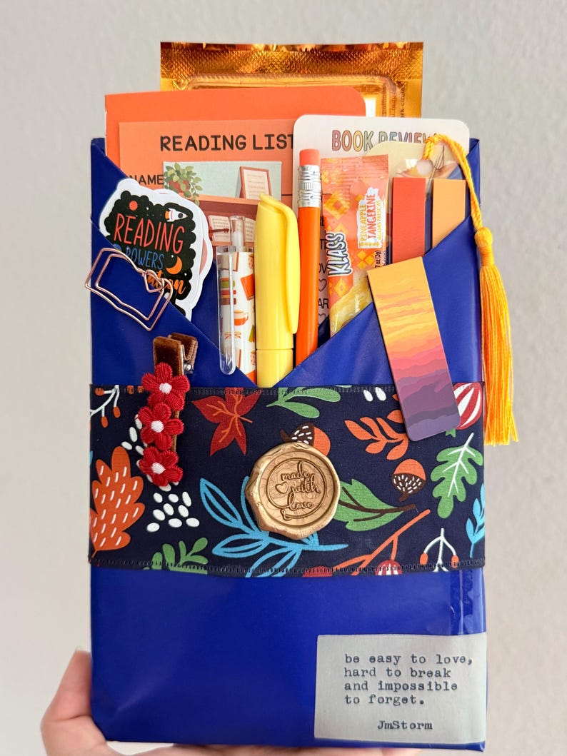 May include: A blue paper-wrapped package containing a reading list, book review card, yellow highlighter, orange pencil, drink mix packet, and a sunset-themed bookmark. It features a floral ribbon, a wax seal, and a label with the text be easy to love.
