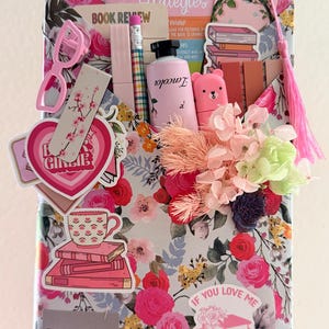 May include: A rectangular gift package wrapped in floral paper featuring pink and red roses. It is decorated with stickers of books, a teacup, and text including Reading Strategies, Book Review, and If You Love Me Read Me A Book. Accessories include a pink tassel, a small bottle, a pencil, and dried flowers.