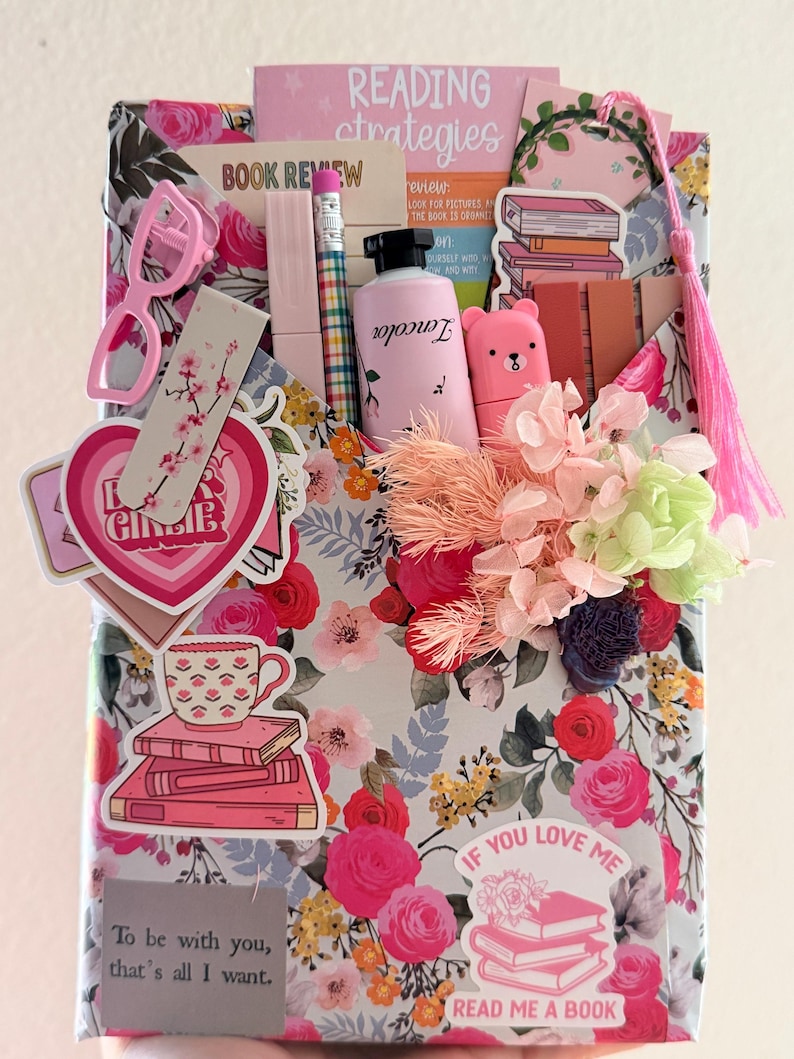 May include: A rectangular gift package wrapped in floral paper featuring pink and red roses. It is decorated with stickers of books, a teacup, and text including Reading Strategies, Book Review, and If You Love Me Read Me A Book. Accessories include a pink tassel, a small bottle, a pencil, and dried flowers.