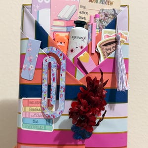 May include: A rectangular paper envelope with horizontal stripes in pink, orange, and dark blue, filled with stationery items including a book review card, a notebook labeled COOL KID, pens, a bear-shaped highlighter, stickers, and a tassel.