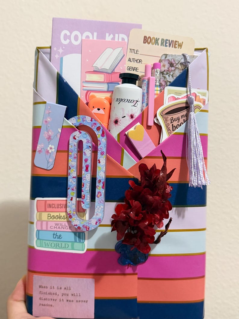 May include: A rectangular paper envelope with horizontal stripes in pink, orange, and dark blue, filled with stationery items including a book review card, a notebook labeled COOL KID, pens, a bear-shaped highlighter, stickers, and a tassel.