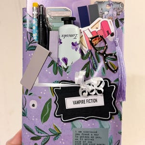 May include: A purple envelope with a green leaf pattern containing stationery items including a pencil, black pen, Zencolor tube, glasses, paper clips, and a label reading VAMPIRE FICTION. A quote by Jessica Katoff is printed on the front.