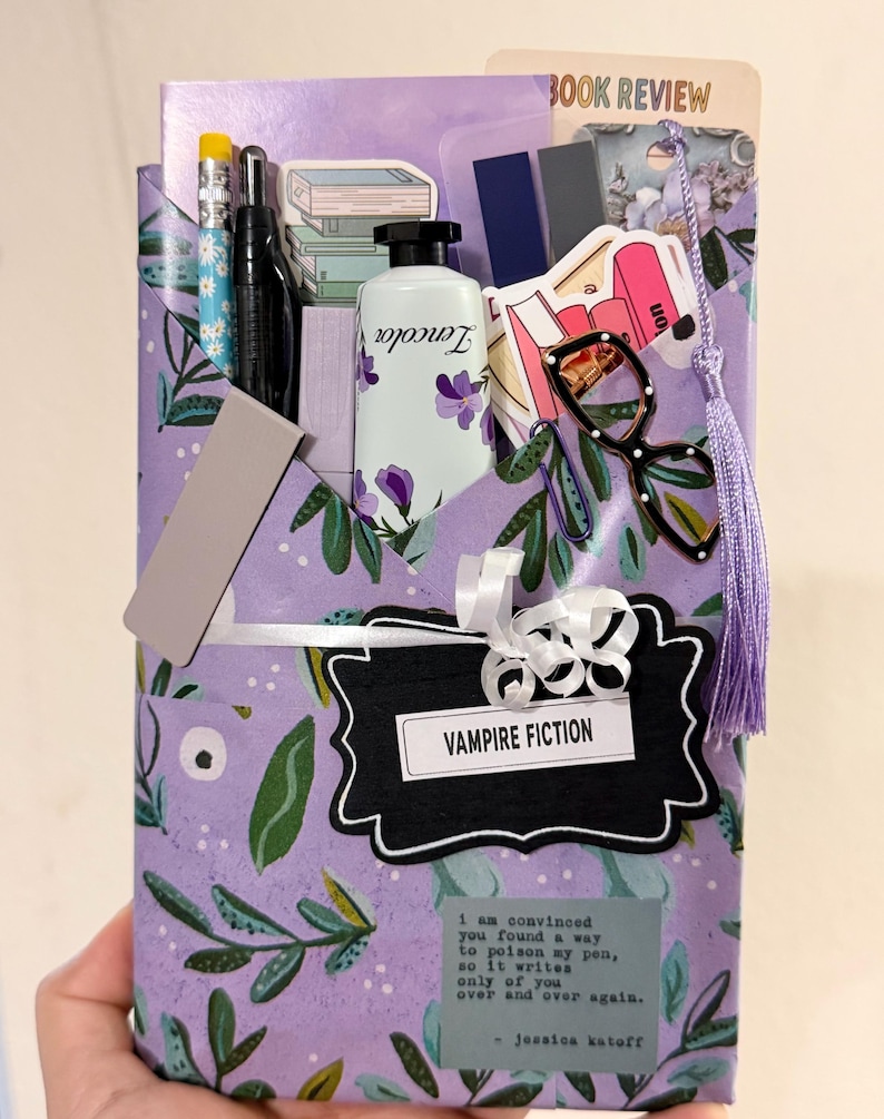 May include: A purple envelope with a green leaf pattern containing stationery items including a pencil, black pen, Zencolor tube, glasses, paper clips, and a label reading VAMPIRE FICTION. A quote by Jessica Katoff is printed on the front.