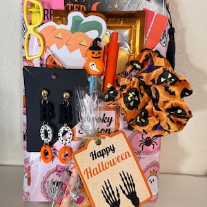 May include: A Halloween-themed gift bag containing a pair of black and white drop earrings, an orange pumpkin-patterned scrunchie, a yellow hair clip, an orange pen with a pumpkin topper, and a tag labeled Happy Halloween with skeleton hands.