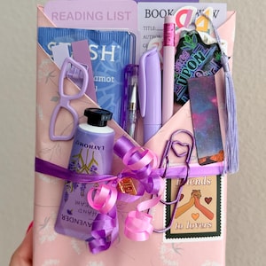 May include: A pink envelope gift set containing a lavender hand cream tube, a purple pen, a pencil, a reading list card, a bookmark with a galaxy print, a heart-shaped paperclip, a small pair of glasses, and a sticker labeled Friends to lovers.