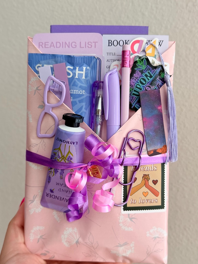 May include: A pink envelope gift set containing a lavender hand cream tube, a purple pen, a pencil, a reading list card, a bookmark with a galaxy print, a heart-shaped paperclip, a small pair of glasses, and a sticker labeled Friends to lovers.