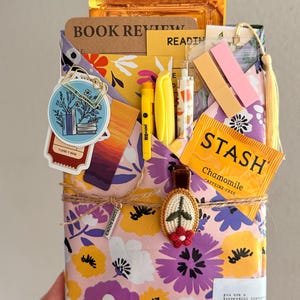 May include: A floral patterned paper envelope containing a book review notebook, a Stash chamomile tea bag, yellow pens, sticky notes, and a circular sticker with a book illustration. The envelope is tied with twine and a small embroidered flower charm.