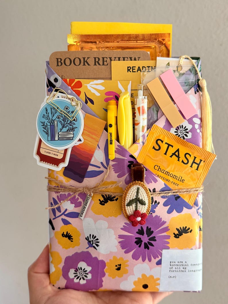 May include: A floral patterned paper envelope containing a book review notebook, a Stash chamomile tea bag, yellow pens, sticky notes, and a circular sticker with a book illustration. The envelope is tied with twine and a small embroidered flower charm.