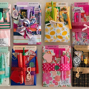 May include: Eight rectangular gift packages arranged in two rows, each wrapped in patterned paper and secured with a ribbon. Each package contains stationery items, pens, reading list cards, and decorative stickers, with some featuring wax seals.