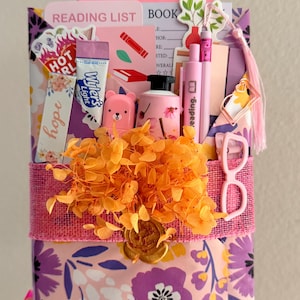 May include: A gift-wrapped book package featuring floral paper in purple, pink, and yellow. It includes a reading list, a Wyler's Light packet, a pink bear figurine, a small bottle, pens, a bookmark, and a pink glasses clip, all adorned with orange dried flowers and a gold wax seal.