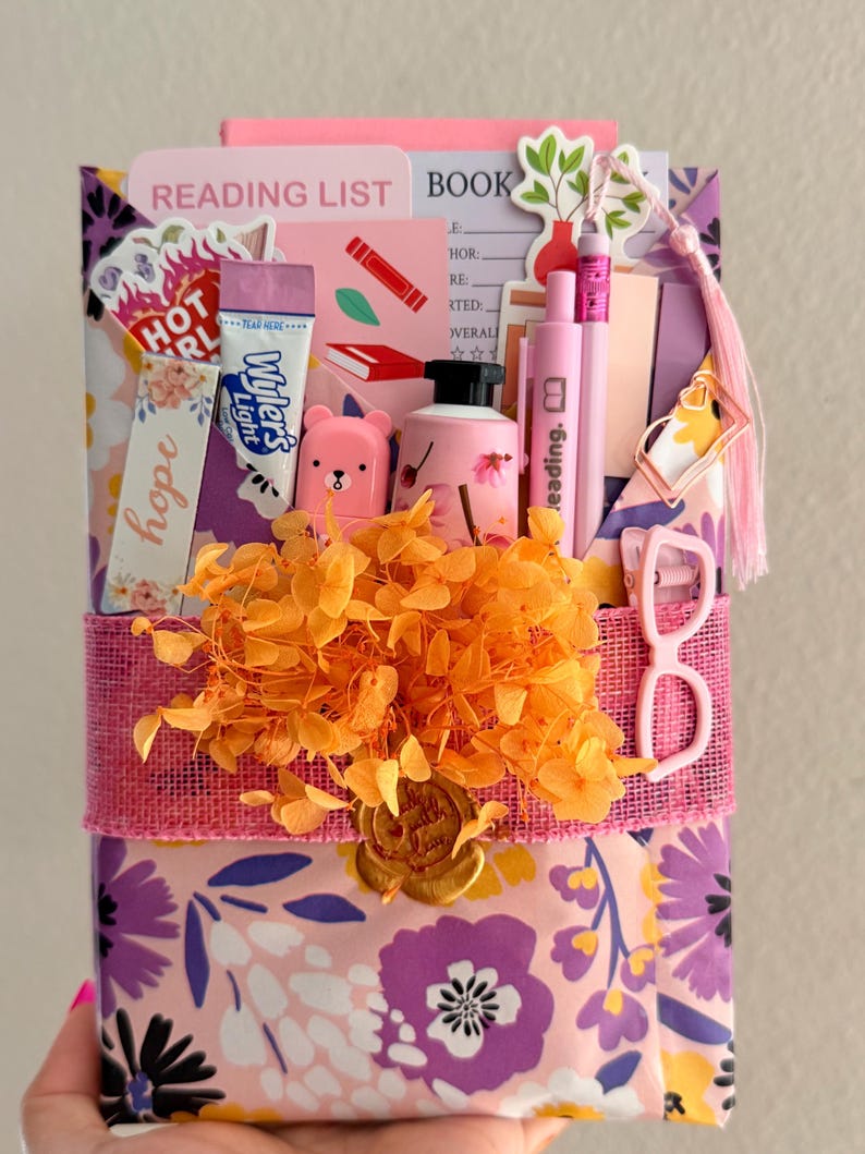 May include: A gift-wrapped book package featuring floral paper in purple, pink, and yellow. It includes a reading list, a Wyler's Light packet, a pink bear figurine, a small bottle, pens, a bookmark, and a pink glasses clip, all adorned with orange dried flowers and a gold wax seal.