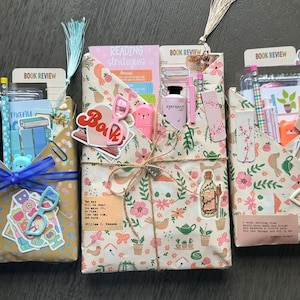 May include: Three rectangular envelopes wrapped in patterned paper, each containing stationery items like pencils, bookmarks, and stickers. The envelopes are decorated with floral prints, text, and small charms, tied with twine or ribbon, and topped with decorative tassels.