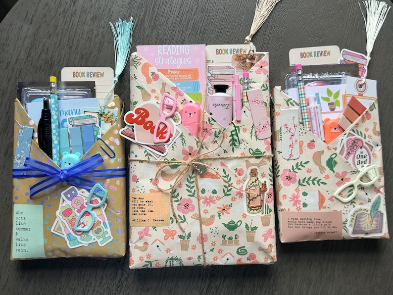 May include: Three rectangular envelopes wrapped in patterned paper, each containing stationery items like pencils, bookmarks, and stickers. The envelopes are decorated with floral prints, text, and small charms, tied with twine or ribbon, and topped with decorative tassels.