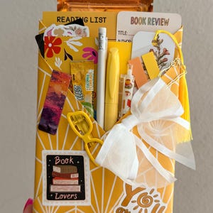 May include: A yellow envelope with a white geometric pattern containing a reading list, book review card, pencil, yellow pen, bookmark, and a white bow. Stickers include a stack of books labeled Book Lovers and text reading You are my sunshine.