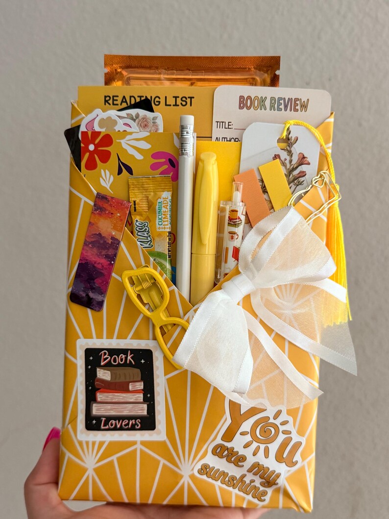 May include: A yellow envelope with a white geometric pattern containing a reading list, book review card, pencil, yellow pen, bookmark, and a white bow. Stickers include a stack of books labeled Book Lovers and text reading You are my sunshine.