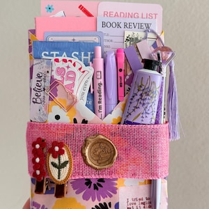 May include: A stationery gift set featuring a floral patterned envelope secured with a pink burlap band, a wax seal, and two embroidered flower clips. It includes a reading list card, a lavender-themed tube, pens, and various paper ephemera.