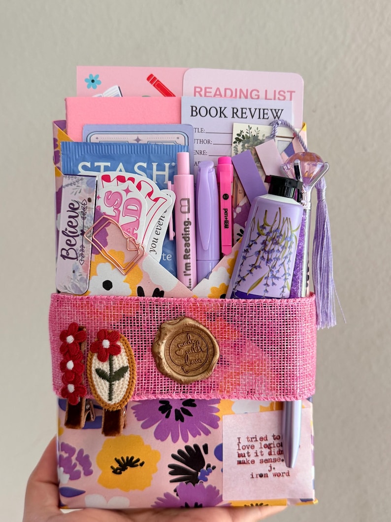 May include: A stationery gift set featuring a floral patterned envelope secured with a pink burlap band, a wax seal, and two embroidered flower clips. It includes a reading list card, a lavender-themed tube, pens, and various paper ephemera.