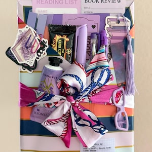 May include: A rectangular gift package wrapped in striped paper featuring a lavender, orange, and navy color palette. The package is adorned with a silk scarf tied in a bow, a lavender hand cream tube, pens, stickers, a bookmark, and paper cards.