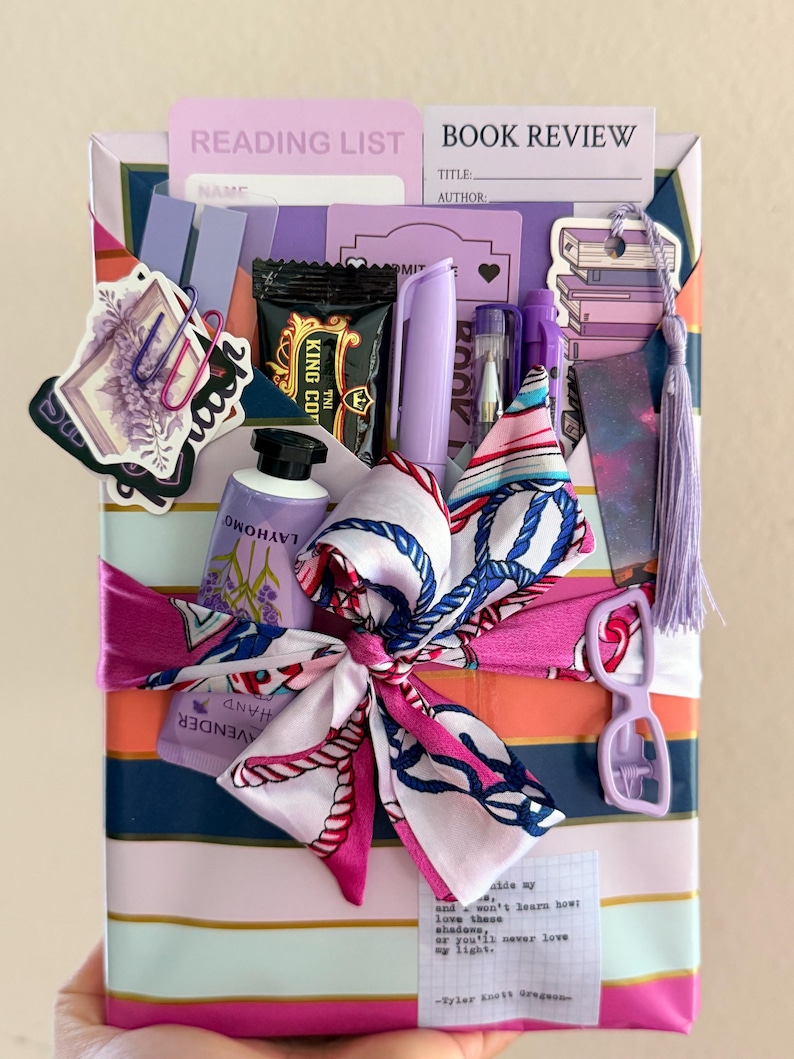 May include: A rectangular gift package wrapped in striped paper featuring a lavender, orange, and navy color palette. The package is adorned with a silk scarf tied in a bow, a lavender hand cream tube, pens, stickers, a bookmark, and paper cards.