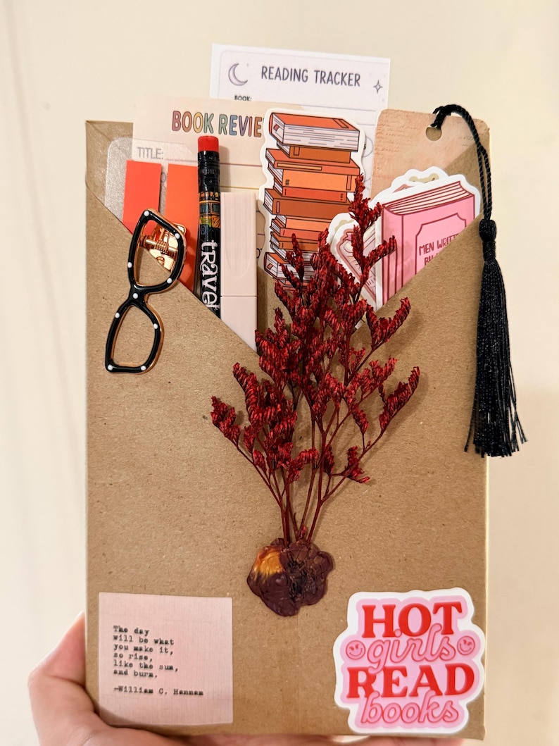 May include: A brown paper envelope containing a reading tracker, book review card, pencil, stickers of books, and a bookmark with a black tassel. A pair of black glasses-shaped pin, a sprig of red dried flowers, and a sticker reading "HOT girls READ books" are attached to the front, along with a quote by William C. Hannan.