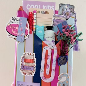 May include: A gift-wrapped package featuring vertical stripes in blue, pink, and gold. It is decorated with various stickers including a stack of books, a door hanger reading GO AWAY, I'M READING, and a bookmark. It contains a pen, pencil, and dried flowers.