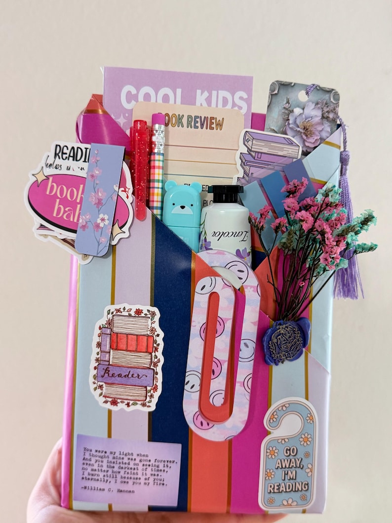 May include: A gift-wrapped package featuring vertical stripes in blue, pink, and gold. It is decorated with various stickers including a stack of books, a door hanger reading GO AWAY, I'M READING, and a bookmark. It contains a pen, pencil, and dried flowers.