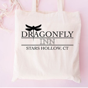May include: A white canvas tote bag with black text and a dragonfly graphic. The text reads "Dragonfly Inn" and "Stars Hollow, CT".