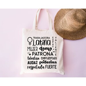 May include: A white tote bag with black text that reads "Trabajadora, Latina, Mujer, Dama, Patrona, Talentosa, Empoderada, Audaz, Poderosa, Respetada, Fuerte". The text is arranged in a circle around a female symbol.
