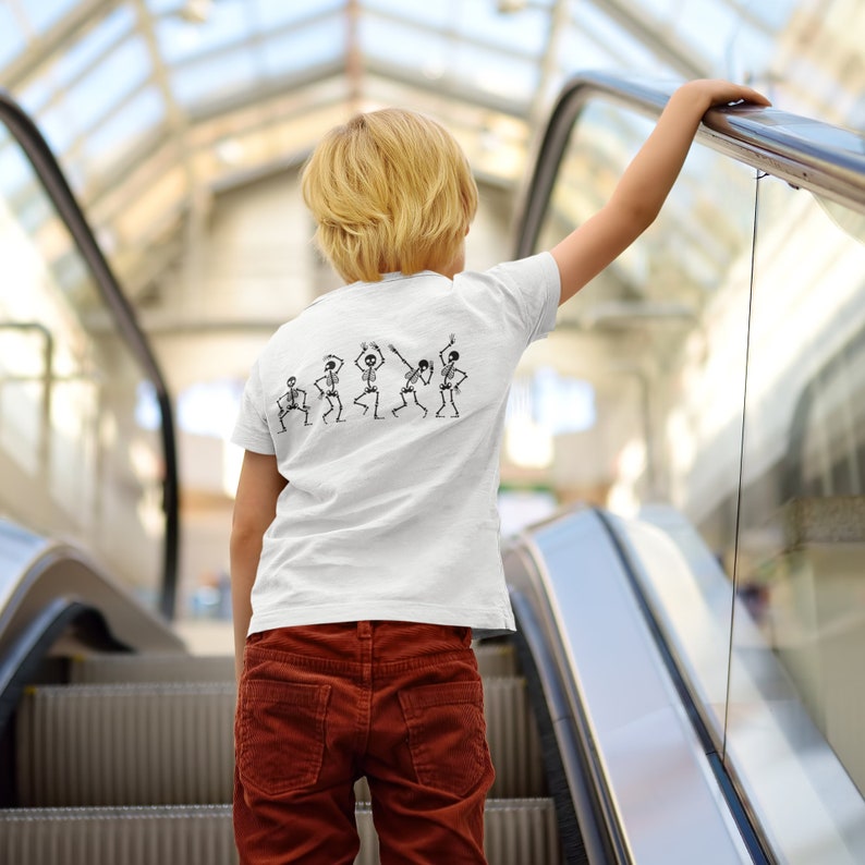 May include: A young person wearing a white t-shirt with a line of dancing skeletons printed on the back. The person is standing on an escalator and holding onto the handrail.
