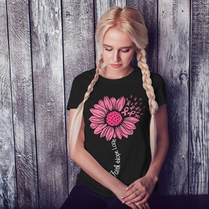 May include: Black t-shirt with a pink sunflower design. The sunflower is made up of pink ribbon symbols. The text "Faith Hope Love" is written vertically down the stem of the sunflower.