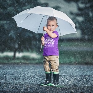May include: A young child wearing a purple shirt with a rainbow and the text "Purple Military Kids" and a heart. The child is holding a white umbrella and standing in the rain.
