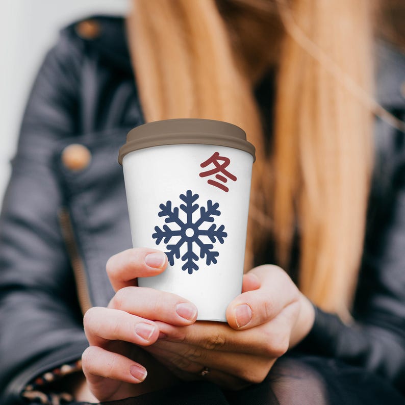 May include: A white paper coffee cup with a brown lid. The cup features a blue snowflake design and red and black Japanese characters. The cup is held by a person with long blonde hair.