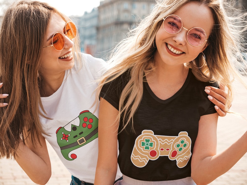 May include: Two friends wearing t-shirts with a Christmas-themed video game controller design. One shirt is white with a green and red controller, the other is black with a brown gingerbread controller.