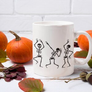 May include: White ceramic mug with a black design of three skeletons dancing. The mug is surrounded by fall leaves and pumpkins.