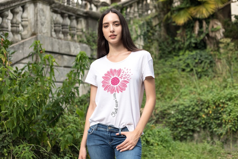 May include: White t-shirt with a pink sunflower design. The sunflower is made up of pink ribbon symbols. The text "Faith Hope Love" is written vertically on the stem of the sunflower.
