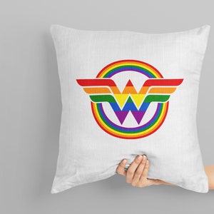 May include: A white pillow featuring a rainbow Wonder Woman logo. The logo is a circle with a rainbow design and a purple W at its centre. The wings are red, orange, yellow, green, blue, and purple.